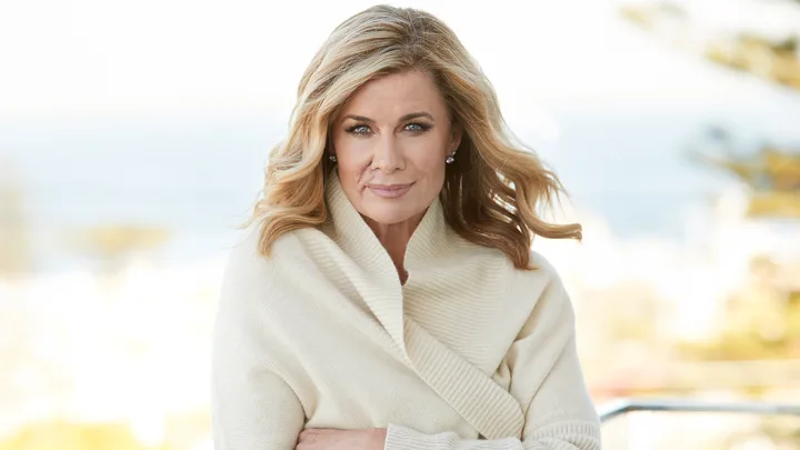 A woman with long blonde hair wearing a cream sweater stands outdoors, smiling softly at the camera.