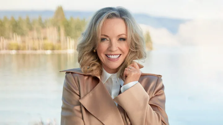 Smiling woman in a tan coat standing near a lake with trees in the background.