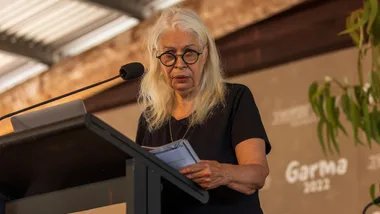 A woman with long white hair and round glasses speaks at a podium with "Garma" banner in the background.