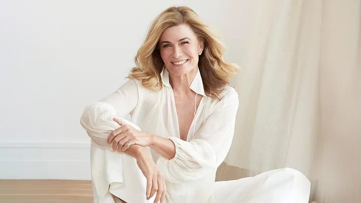 Smiling woman with blonde hair wearing a white outfit, sitting indoors against a light background.