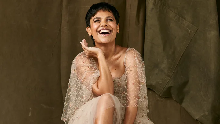 A person with short hair laughs joyfully, wearing a sheer, embroidered dress, seated against a dark fabric background.