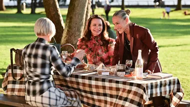 Three women enjoy a picnic at a checkered table in a sunny park setting.