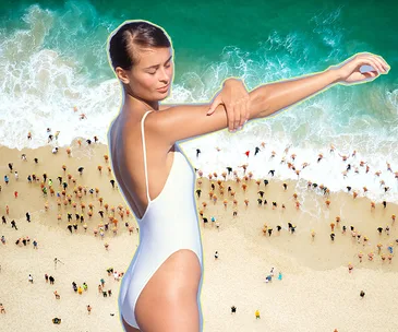 A woman in a white swimsuit applies sunscreen at the beach, with a lively shoreline and turquoise waves in the background.