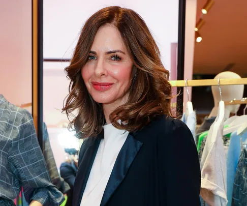 A woman with shoulder-length brown hair, wearing a dark blazer and white blouse, smiles at the camera in a clothing store.