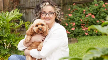 Woman with glasses hugging a small, curly-haired dog in a garden with pink flowers.