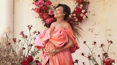 A woman in a vibrant pink dress poses joyfully in front of floral arrangements.