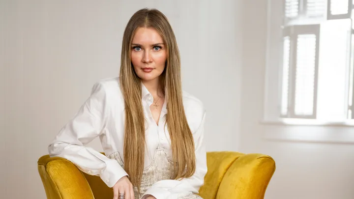 A woman with long hair sits on a yellow chair, wearing a white blouse, in a bright room.