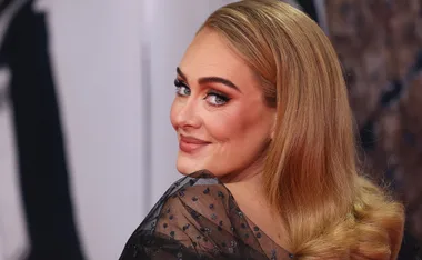 A woman with long blonde hair in a sheer black dress, smiling and looking over her shoulder at the camera.