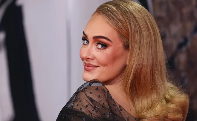 A woman with long blonde hair in a sheer black dress, smiling and looking over her shoulder at the camera.