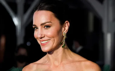 A smiling woman in a green dress, wearing an ornate gold earring, stands in a dimly lit setting.