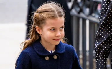 Girl with long hair and navy coat smiling slightly, looking to her right, against an outdoor railing.