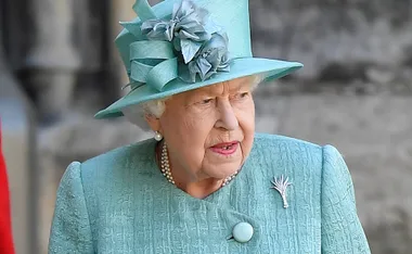 Elderly woman in a teal dress and hat with floral decoration, wearing pearls, looking to the side.