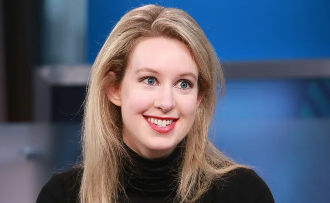 Smiling woman with long blonde hair in a black top, against a blue background.