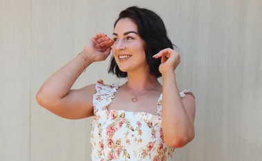 Woman smiling in a floral dress, posing with hands near her face against a light background.