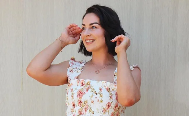 Woman smiling in a floral dress, posing with hands near her face against a light background.
