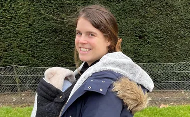 A woman in a blue coat with fur trim smiles while holding a baby, standing in front of a hedge and wire fence.