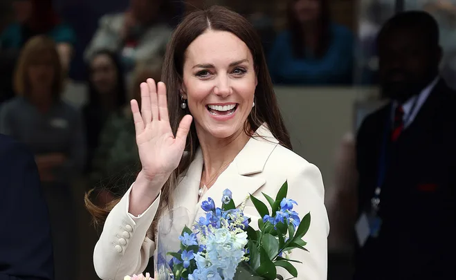 A woman in a white coat waves and smiles, holding a bouquet of blue flowers.