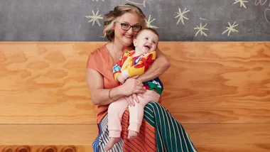Woman with glasses cheerfully holding a smiling baby, both sitting on a wooden bench against a chalkboard background.