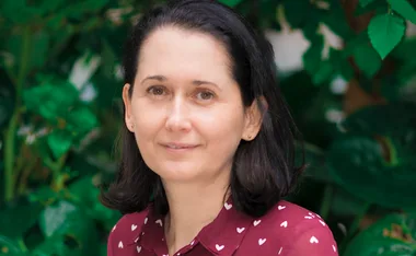 Portrait of a woman with dark hair wearing a maroon shirt, standing against a leafy green background.