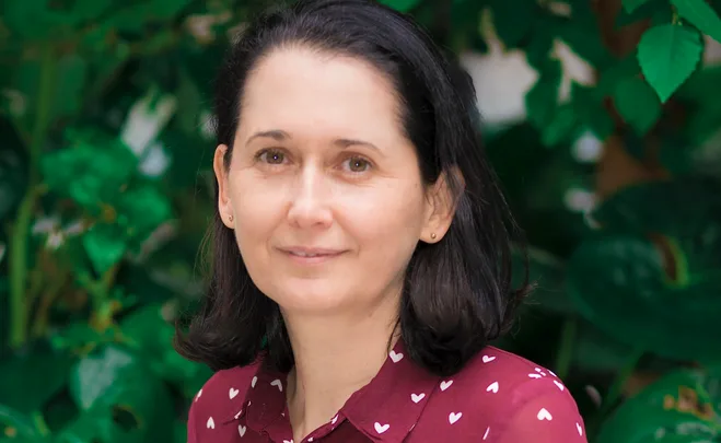 Portrait of a woman with dark hair wearing a maroon shirt, standing against a leafy green background.
