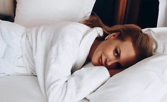 Person in a white robe lying on a bed, smiling at the camera.