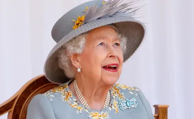 An elderly woman in a floral dress and hat, smiling while looking upwards.