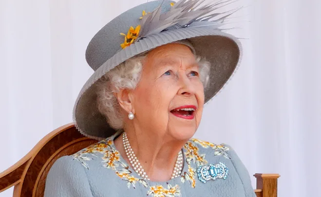 An elderly woman in a floral dress and hat, smiling while looking upwards.