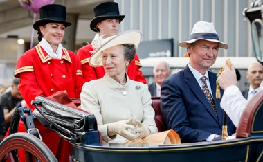 Royal couple in carriage, dressed formally, with attendants in red. Hat and gloves visible, outside urban setting.