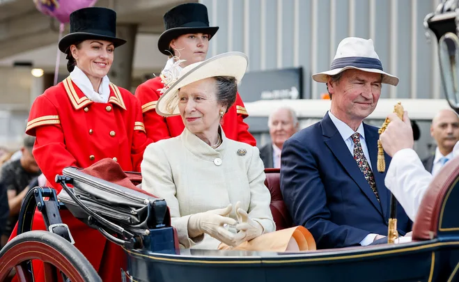 Royal couple in carriage, dressed formally, with attendants in red. Hat and gloves visible, outside urban setting.