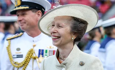 Princess Anne smiling, wearing a cream hat and coat, with a Navy officer in the background during Australia tour 2022.