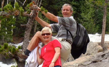 A man and woman smiling outdoors on a rocky terrain with trees in the background.