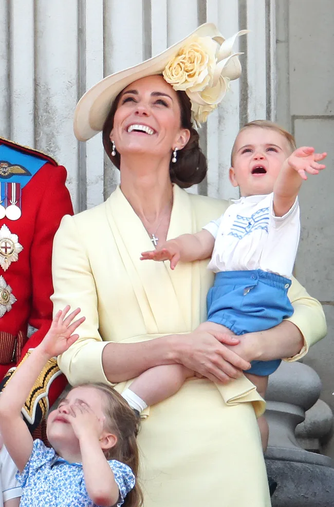 A woman in a yellow outfit and hat holds a young child, both looking up and smiling, with a little girl reaching up beside them.