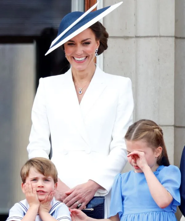 Woman in white with hat smiling, standing with two children; one in blue dress, other in striped shirt with hands on face.