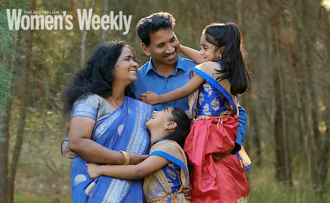 A smiling family embraces outdoors, dressed in traditional attire, with trees in the background.