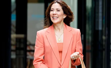 A woman smiling in an orange suit, holding a beige bag, standing outdoors in front of a building.