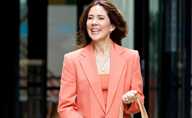 A woman smiling in an orange suit, holding a beige bag, standing outdoors in front of a building.