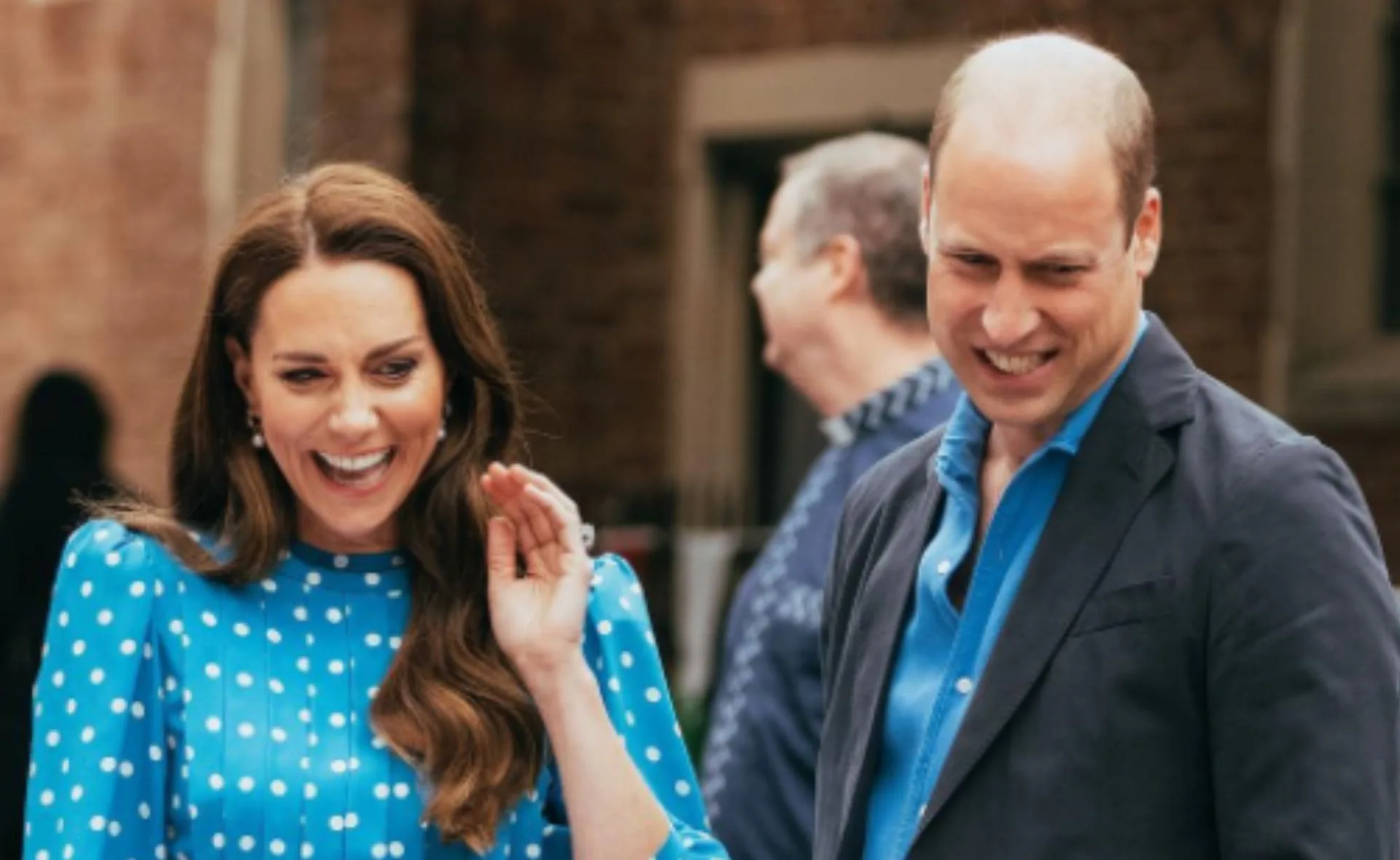 A man and woman smiling and waving outdoors, the woman in a blue polka-dot dress and the man in a dark suit.