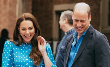 A man and woman smiling and waving outdoors, the woman in a blue polka-dot dress and the man in a dark suit.