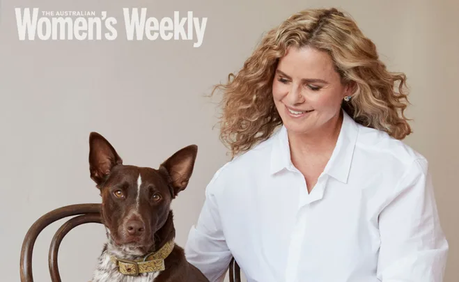 A woman in a white shirt sits smiling beside a brown and white dog on a chair, featured in a magazine cover.