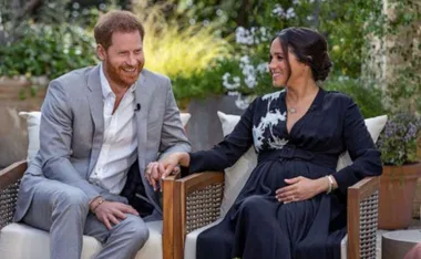 A couple sitting outdoors, holding hands; the man is in a grey suit, the woman in a black dress, smiling at each other.