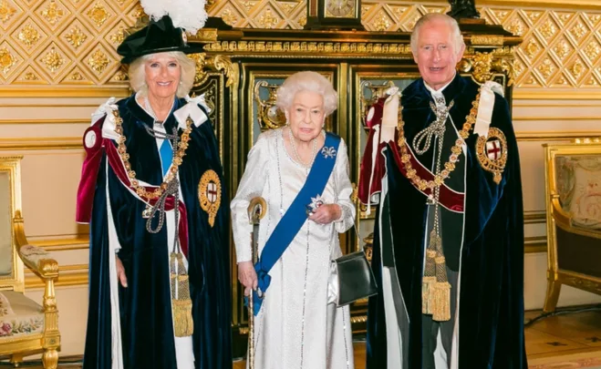 Three people in ceremonial attire, including velvet robes and insignia, stand indoors against a gold-patterned wall.