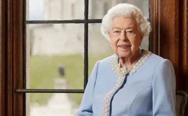Elderly woman with white hair in a light blue dress, seated by a window with blurred background.