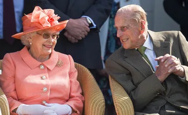 An elderly couple seated outdoors, the woman in a coral outfit and hat, both smiling at each other warmly.