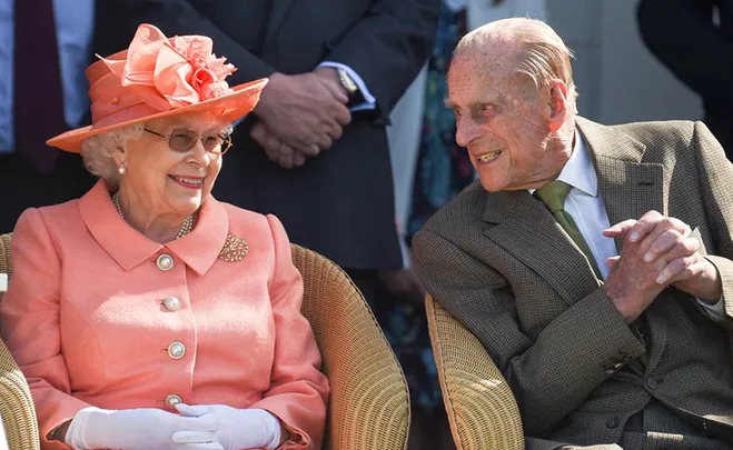 An elderly couple seated outdoors, the woman in a coral outfit and hat, both smiling at each other warmly.
