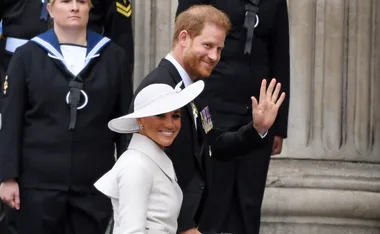 A man in a suit waves, smiling with a woman in a white hat, outside a building, with military personnel in the background.