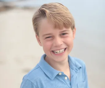 A young boy with blonde hair wearing a blue shirt smiles at the camera against a blurred beach background.