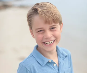 A young boy with blonde hair wearing a blue shirt smiles at the camera against a blurred beach background.