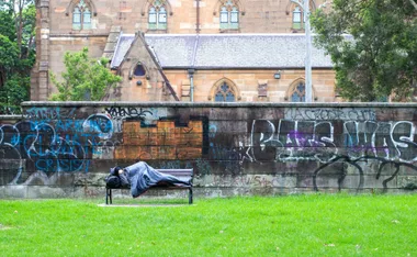 A person sleeps on a park bench with graffiti on the wall behind, near a historic building.
