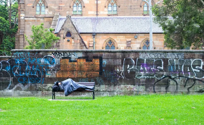 A person sleeps on a park bench with graffiti on the wall behind, near a historic building.