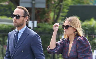 A couple in formal attire and sunglasses walking outdoors, with greenery in the background.